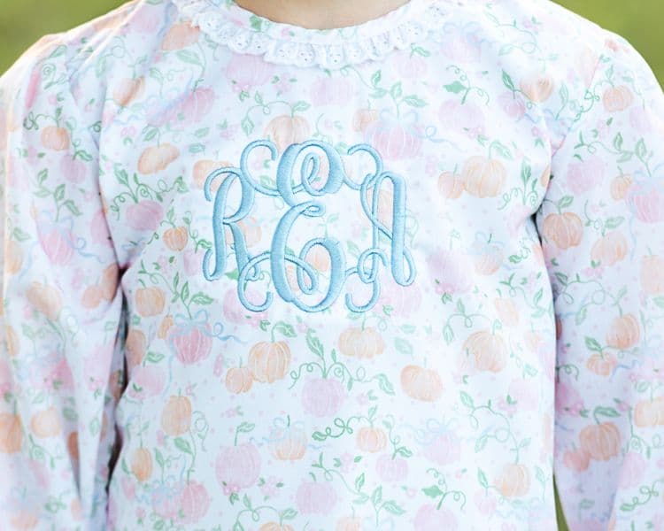 Young girl in a floral blouse with monogram and light blue pants standing in a grassy field.0