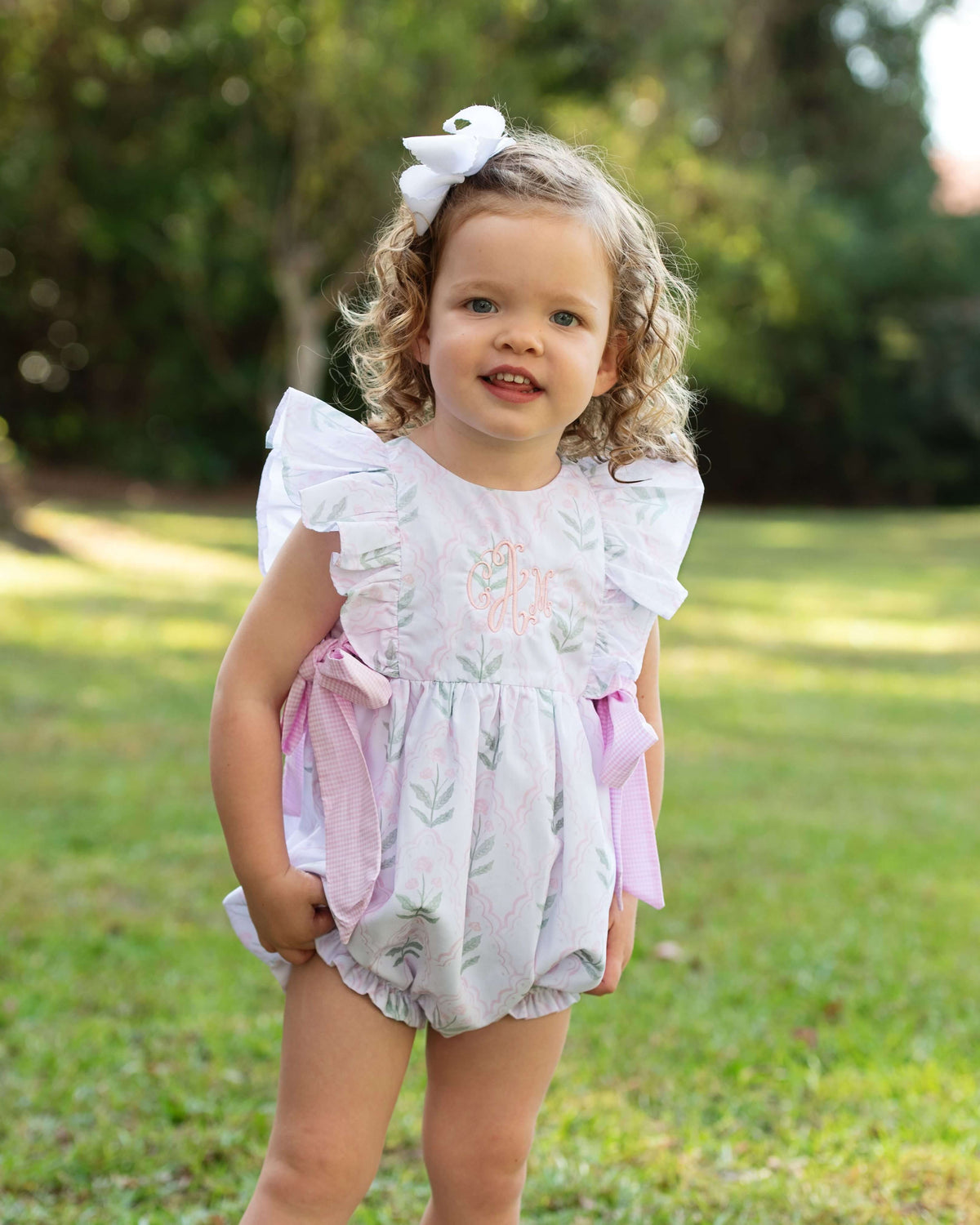 Young girl in a floral bubble with ruffles and bow standing outdoors on grass.