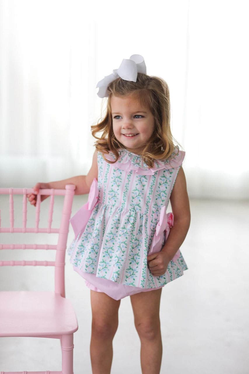Young girl in a floral diaper set with pink trim standing next to a pink chair.