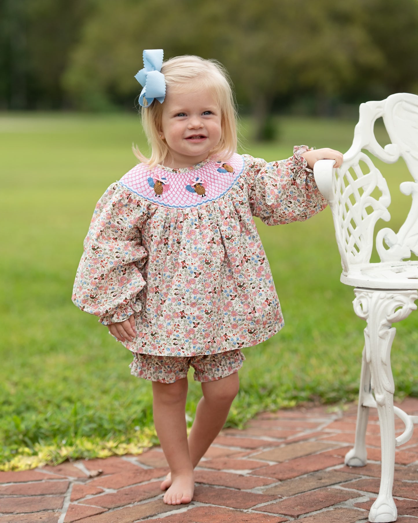 Young girl in a floral diaper set with smocked turkeys and a blue bow standing on a brick path outdoors.