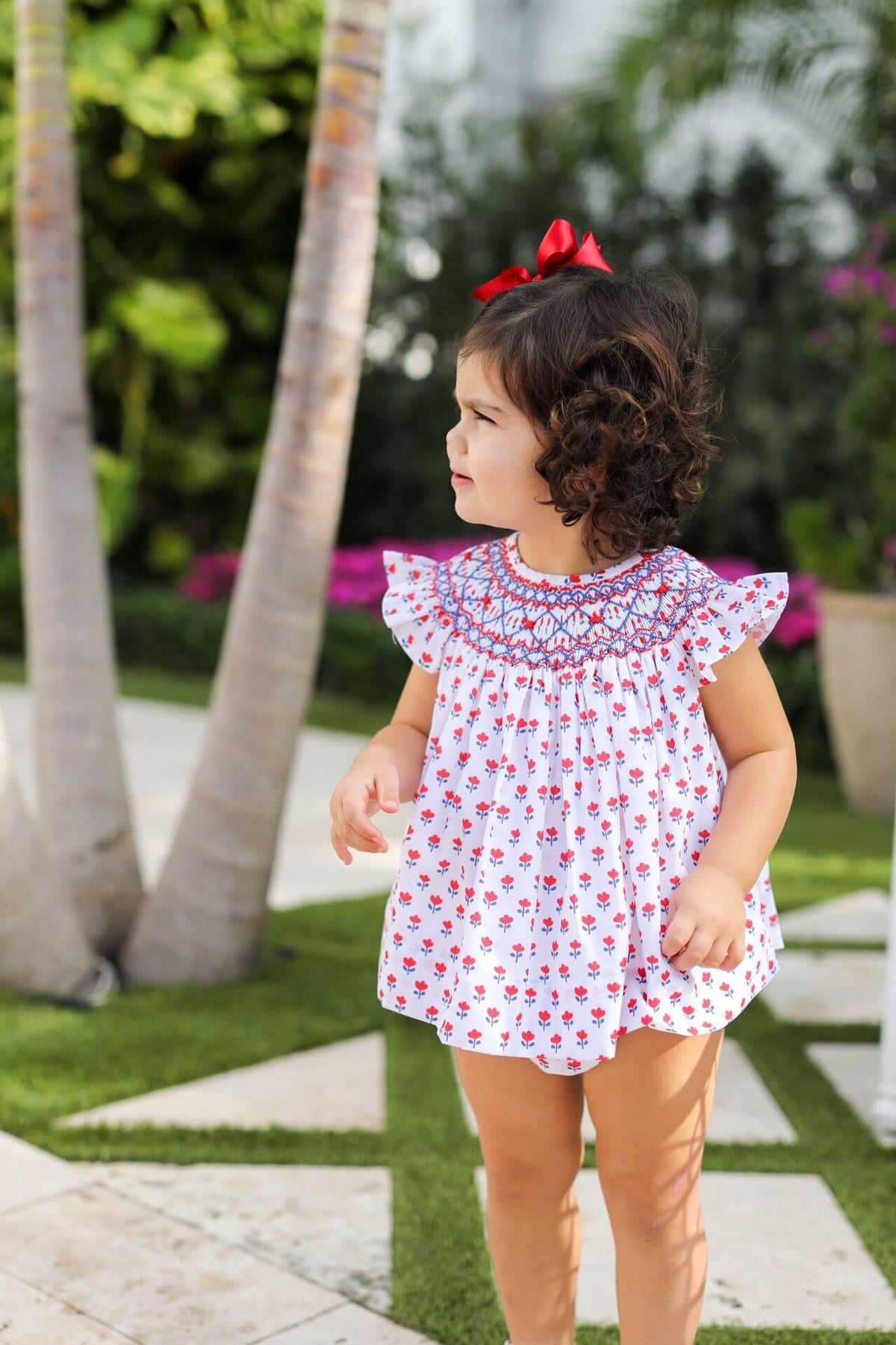 Young girl in a floral dress standing outdoors with palm trees and greenery in the background