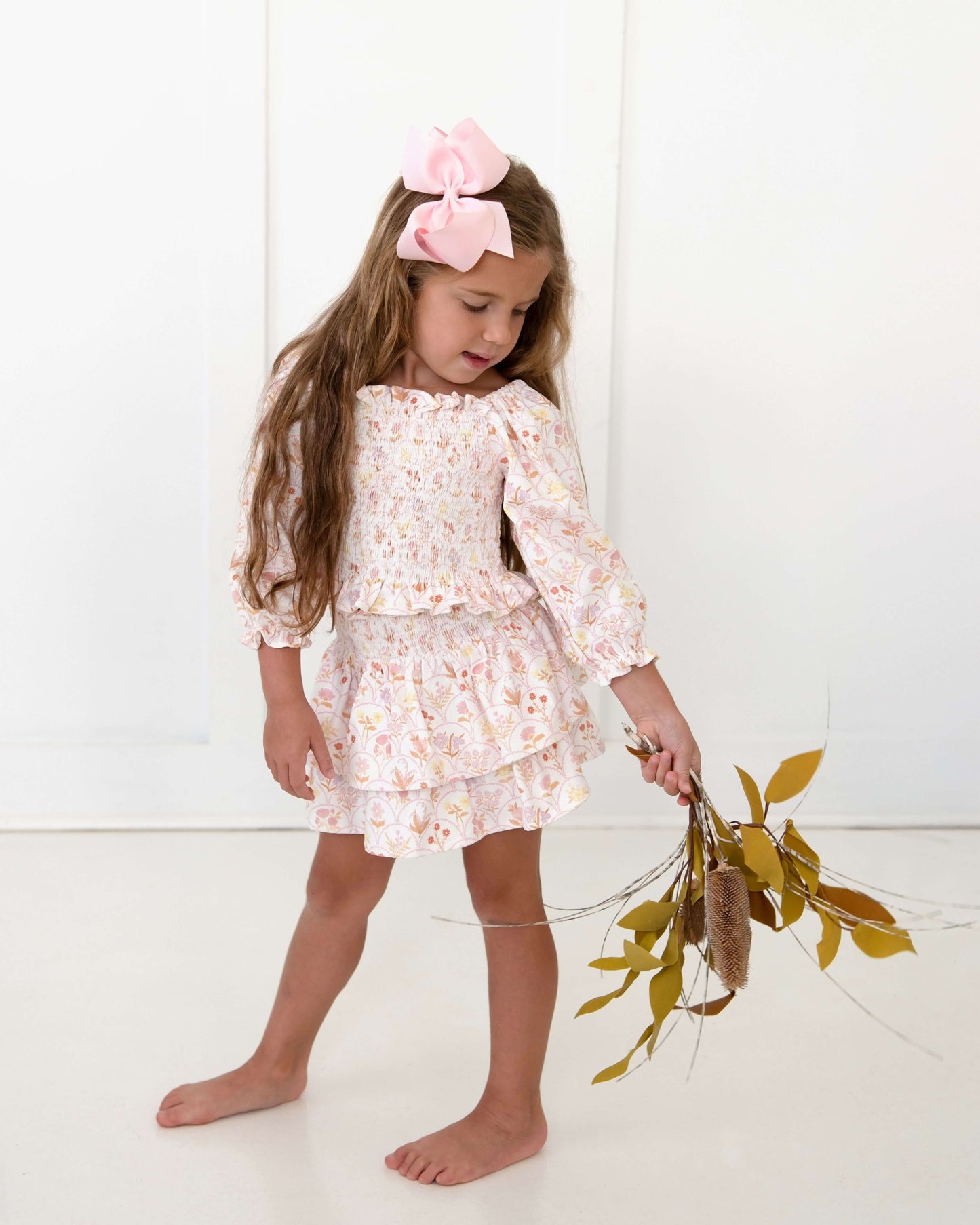 Young girl in a floral dress with a pink bow, holding dried plants against a white background