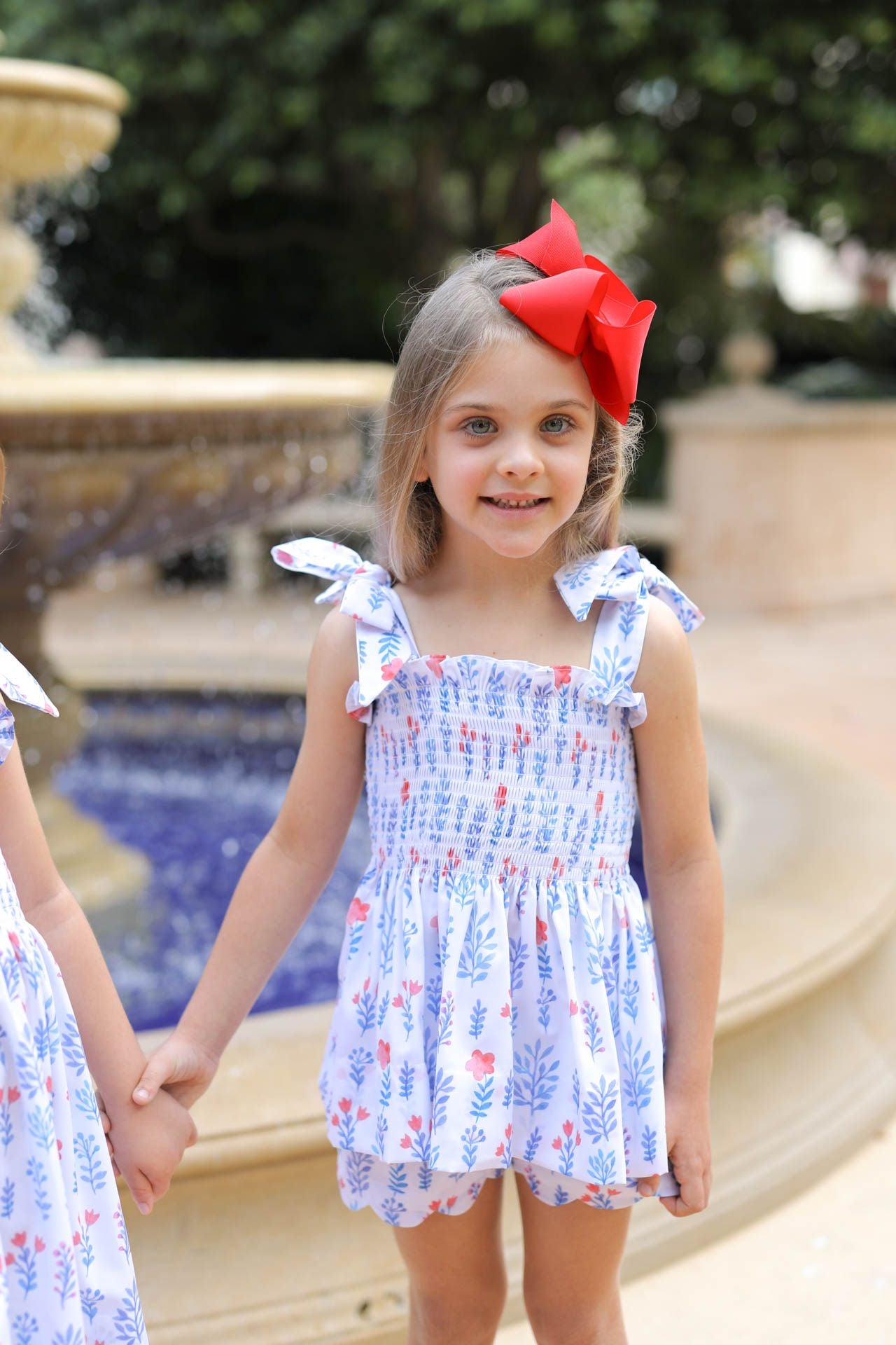 Young girl in a floral dress with a red bow standing in front of a fountain.