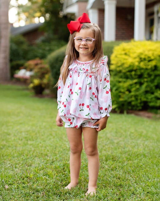 Young girl in a floral dress with cherries and a red bow in her hair, standing on grass.