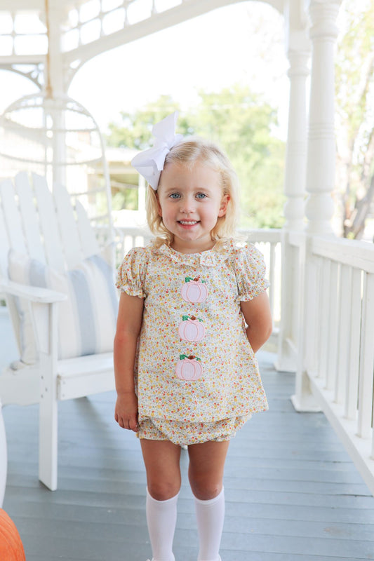 Young girl in a floral dress with pumpkin designs on a white porch.