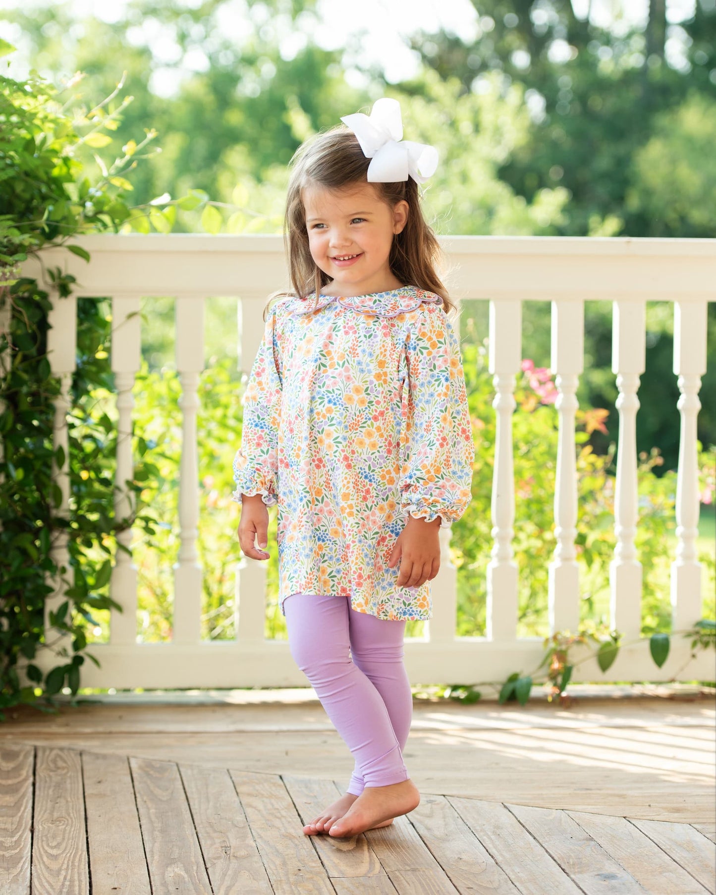 Young girl in a floral legging set standing on a wooden deck with greenery in the background