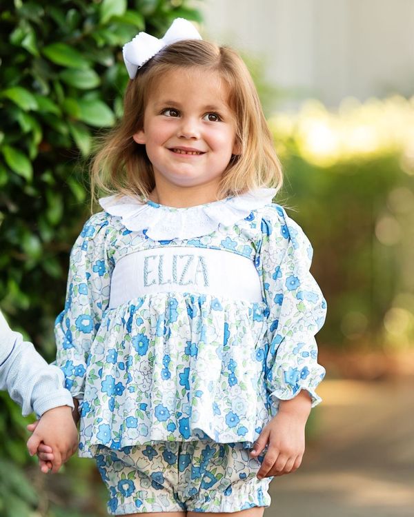 Young girl in a floral outfit with 'Eliza' on it, standing outdoors.