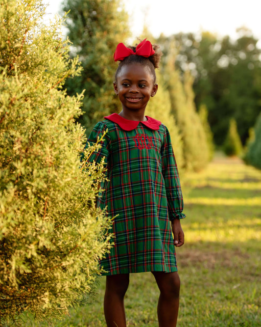 Young girl in a green plaid dress with a red bow standing in a field of trees.