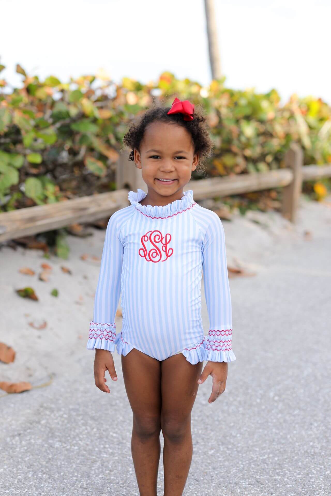 Young girl in a light blue and white striped swimsuit with red monogram, standing on a sandy beach.