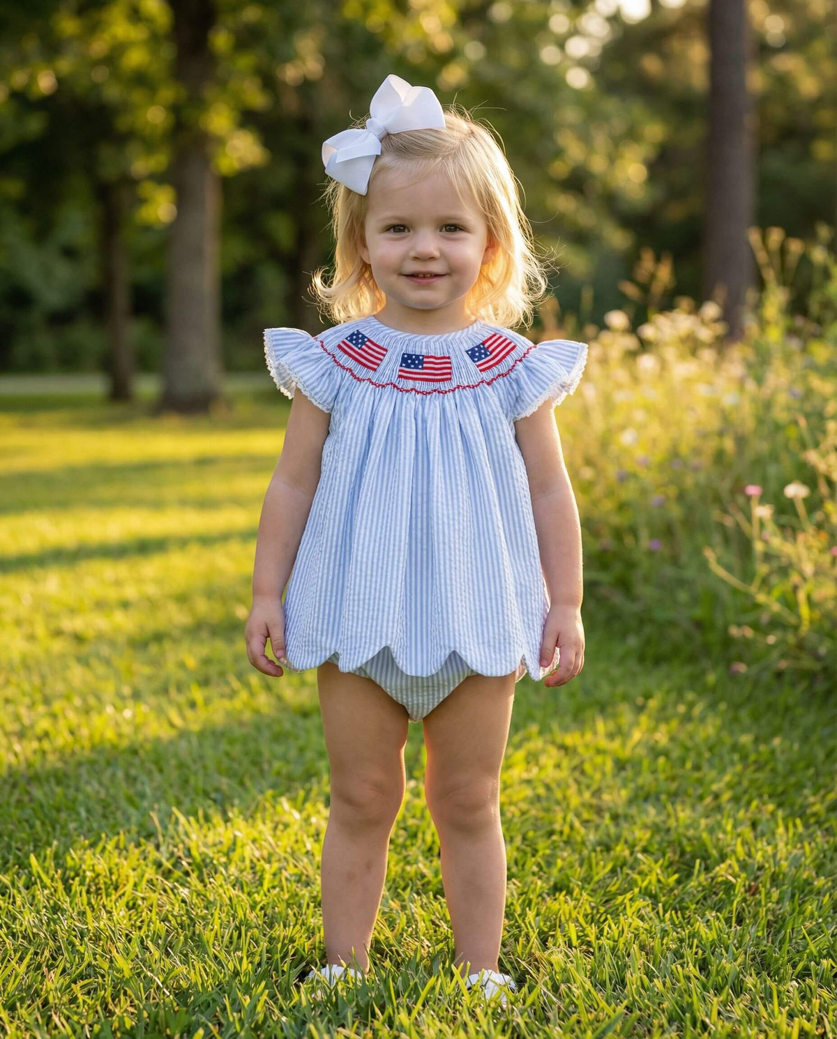 Young girl in a light blue dress with American flag designs standing in a grassy field.