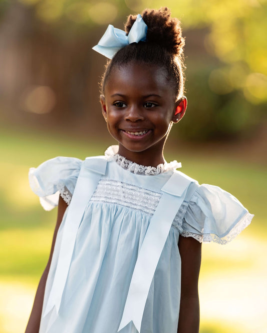 Young girl in a light blue dress with a bow in her hair, standing outdoors.