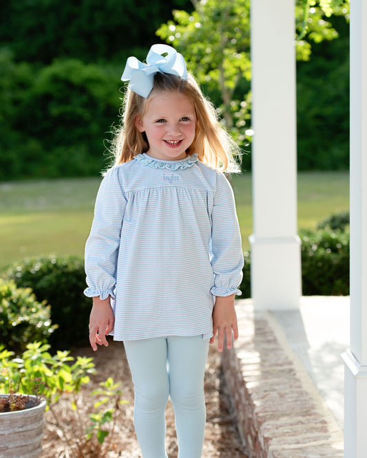 Young girl in a light blue dress with a bow in her hair, standing outdoors.