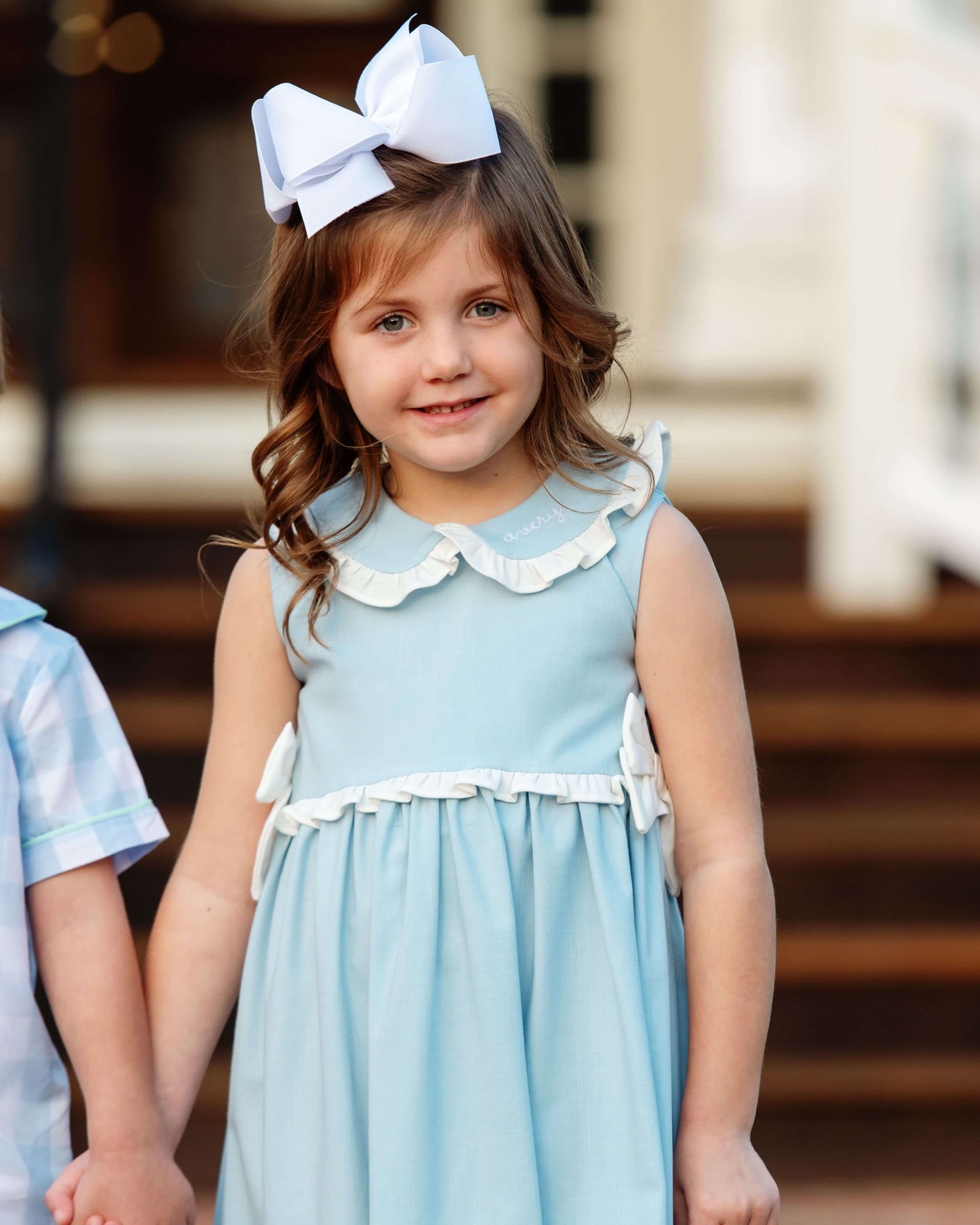 Young girl in a light blue dress with a large bow in her hair, standing outdoors.