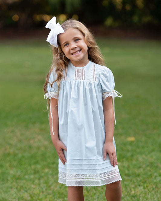 Young girl in a light blue dress with lace details standing on grass.