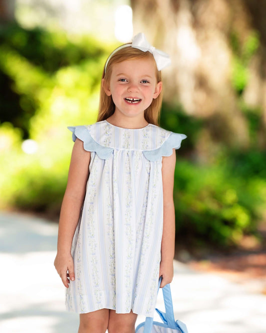 Young girl in a light blue dress with ruffled sleeves standing outdoors with greenery in the background