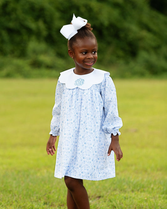 Young girl in a light blue floral dress with a white collar standing in a grassy field.