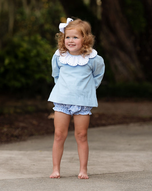 Young girl in a light blue outfit with a white collar standing outdoors.
