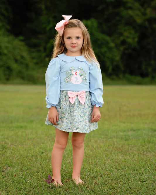 Young girl in a light blue shirt with an applique turkey and floral skirt standing in a grassy field.