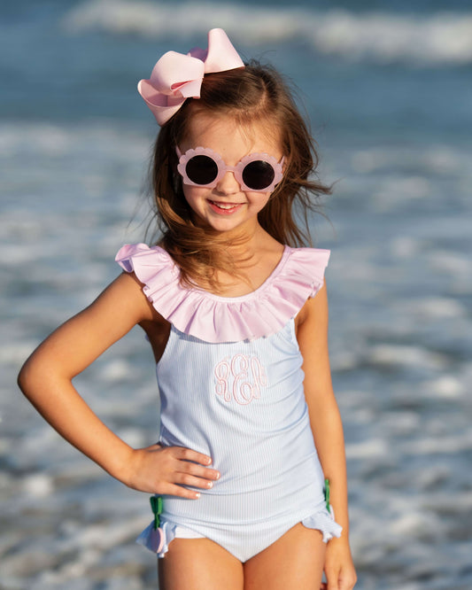 Young girl in a light blue swimsuit with pink ruffled top and sunglasses standing by water.