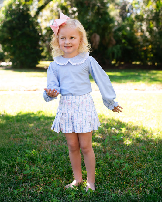 Young girl in a light blue top and white skirt with a pink bow standing in a grassy area.