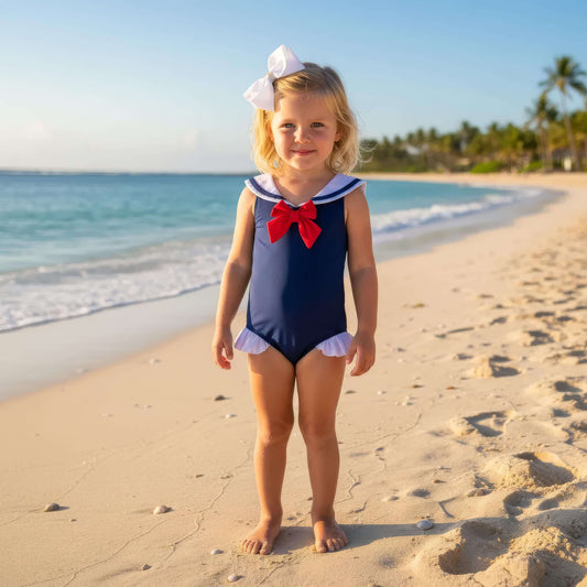 Young girl in a navy blue swimsuit with a red bow standing on a beach.