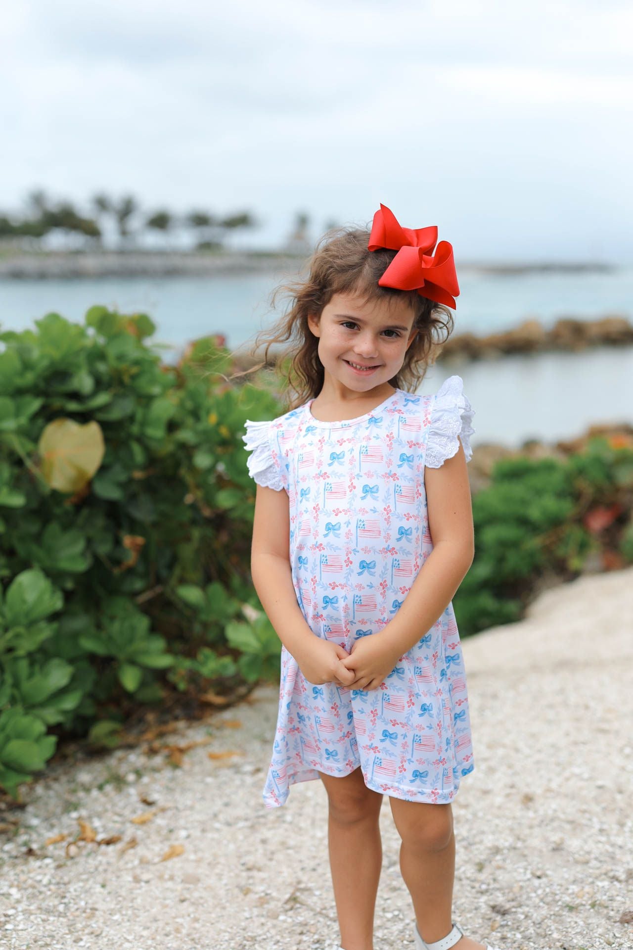 Young girl in a patterned dress with a red bow standing by a waterfront.
