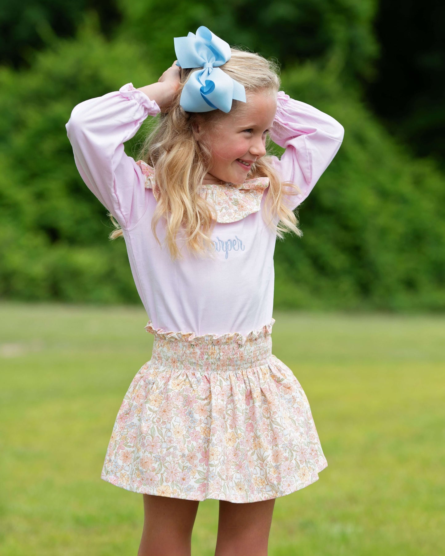 Young girl in a pink blouse and floral skirt with a blue bow in her hair outdoors.