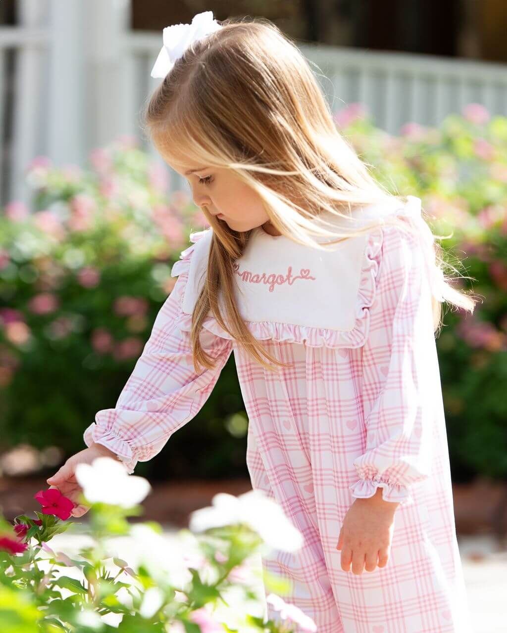 Young girl in a pink checkered dress with a white bow in her hair, standing in a garden.