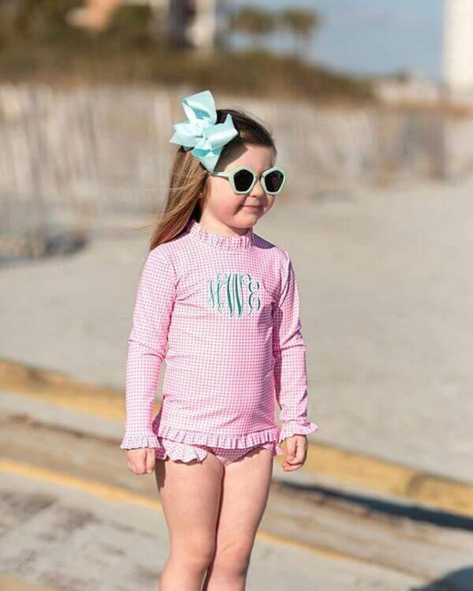 Young girl in a pink checkered swimsuit with sunglasses and a bow on a beach.