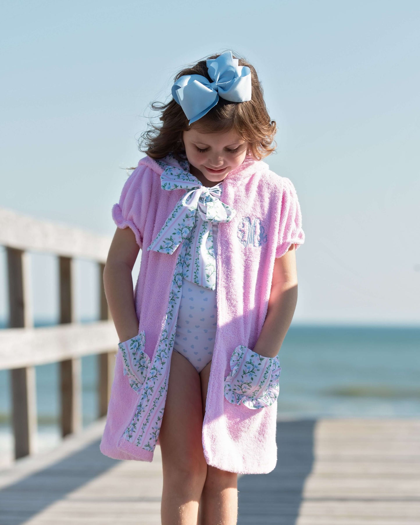 Young girl in a pink cover-up with floral details standing on a wooden pier by the ocean.
