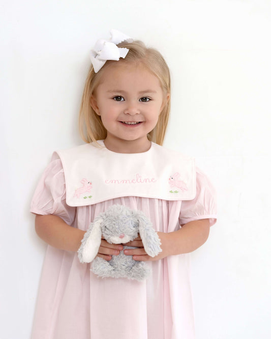 Young girl in a pink dress with a white bib holding a gray teddy bear against a white background