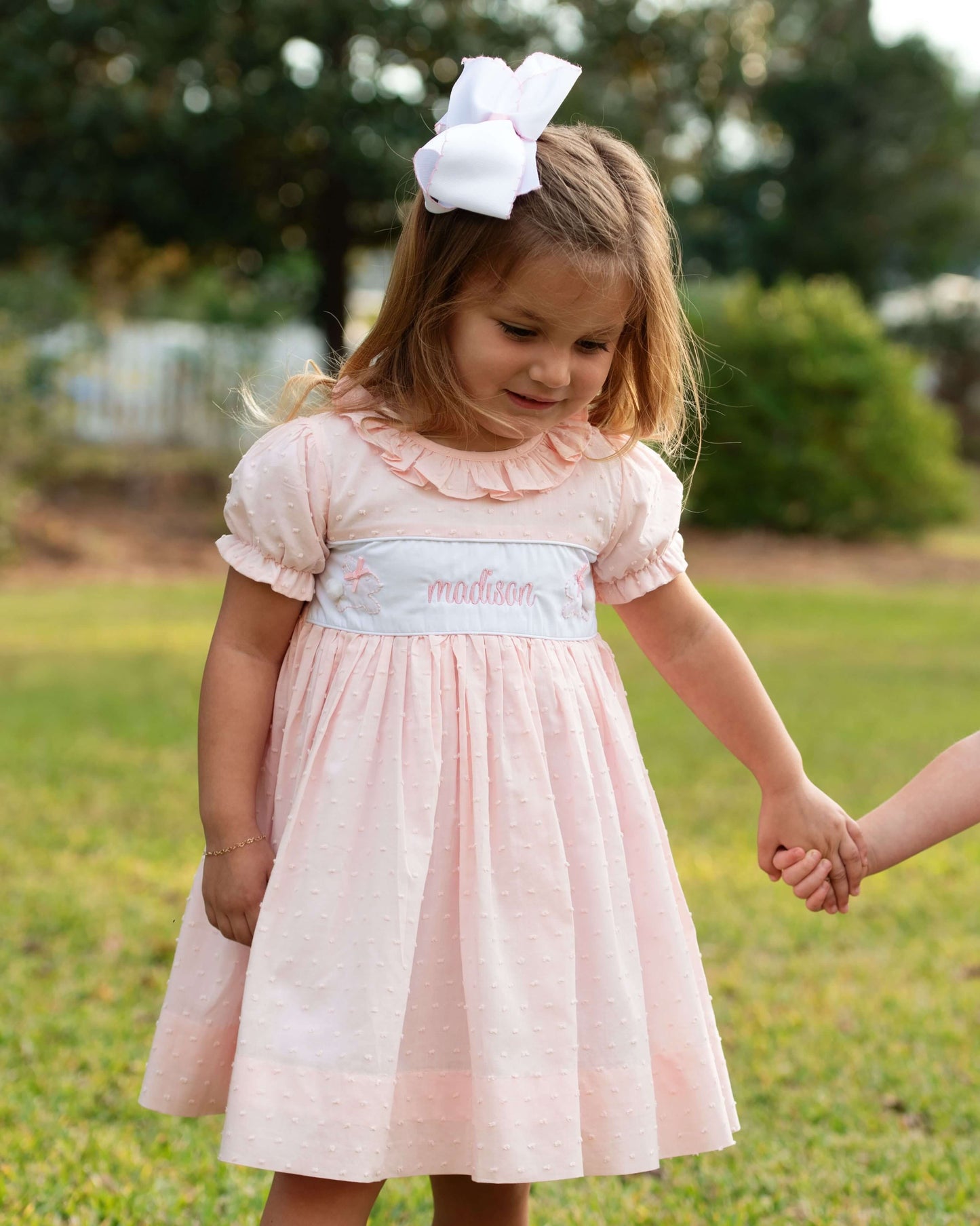 Young girl in a pink dress with a white bow standing outdoors on grass.