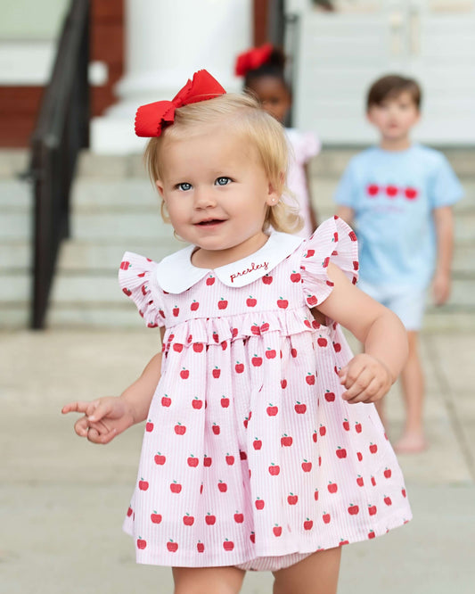 Young girl in a pink dress with red polka dots and a large red bow, standing on a sidewalk.