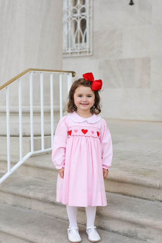 Young girl in a pink dress with red smocked hearts standing on steps outdoors.