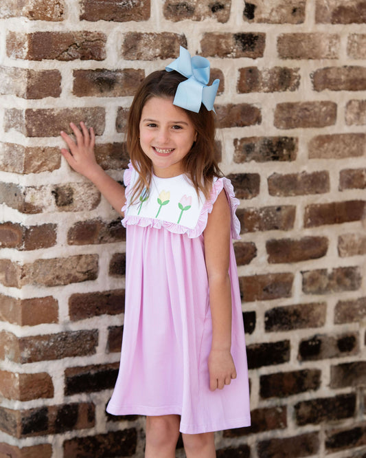 Young girl in a pink dress with white ruffled collar standing against a brick wall.