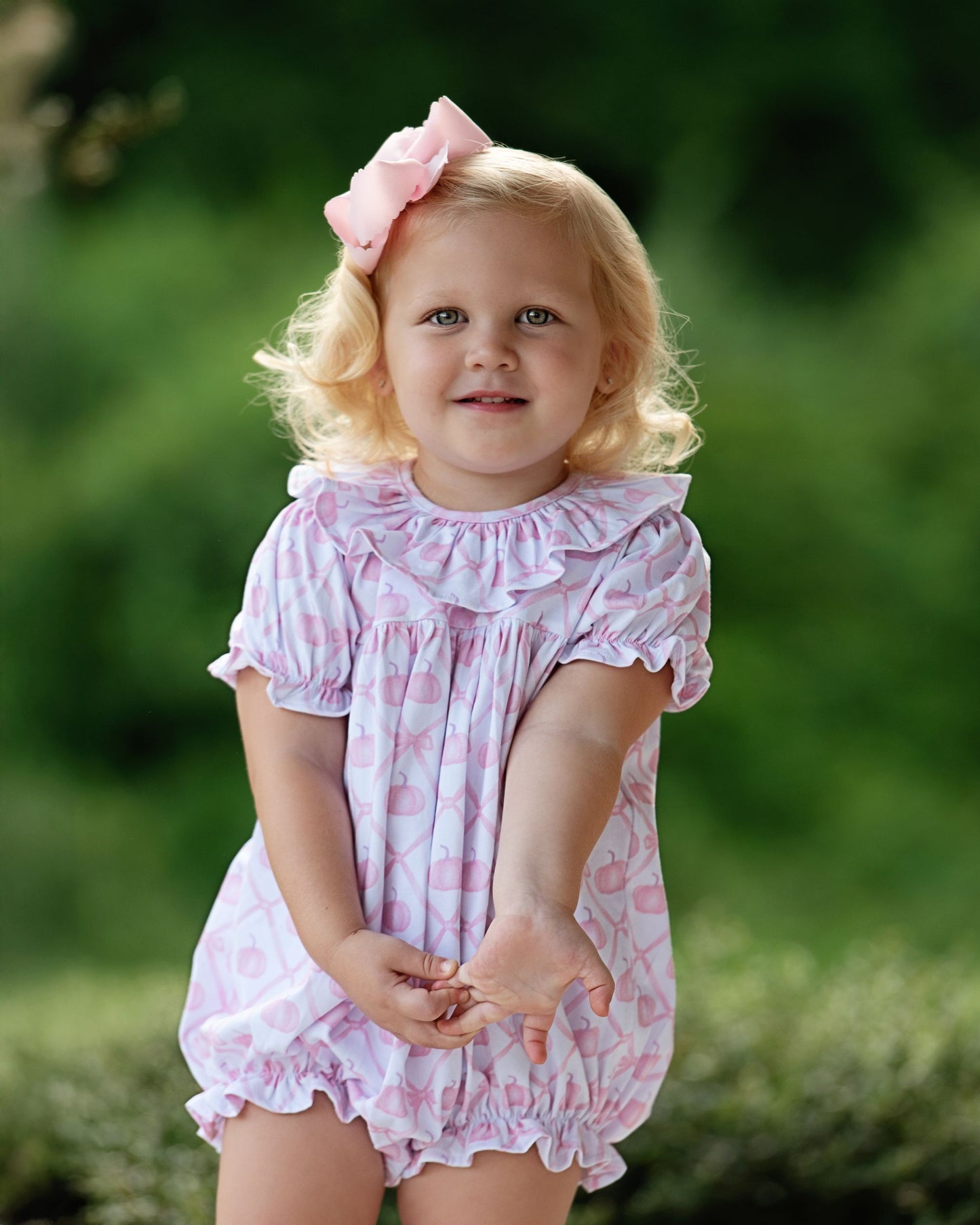 Young girl in a pink floral dress with a bow in her hair, standing outdoors.