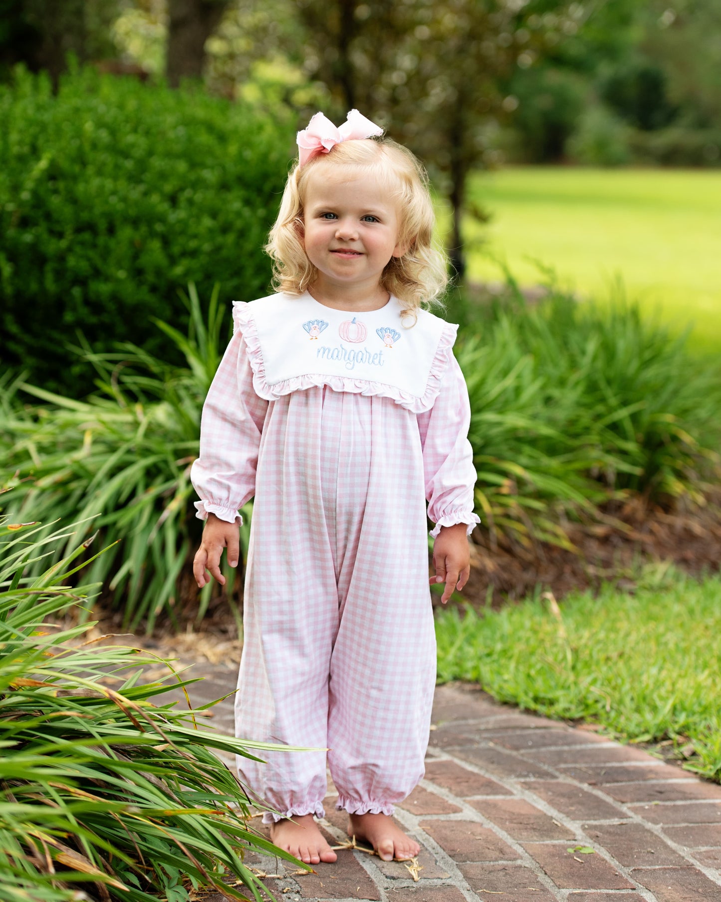 Young girl wearing pink check long bubble with embroidered turkeys and a pumpkin standing on a brick path with greenery around her.