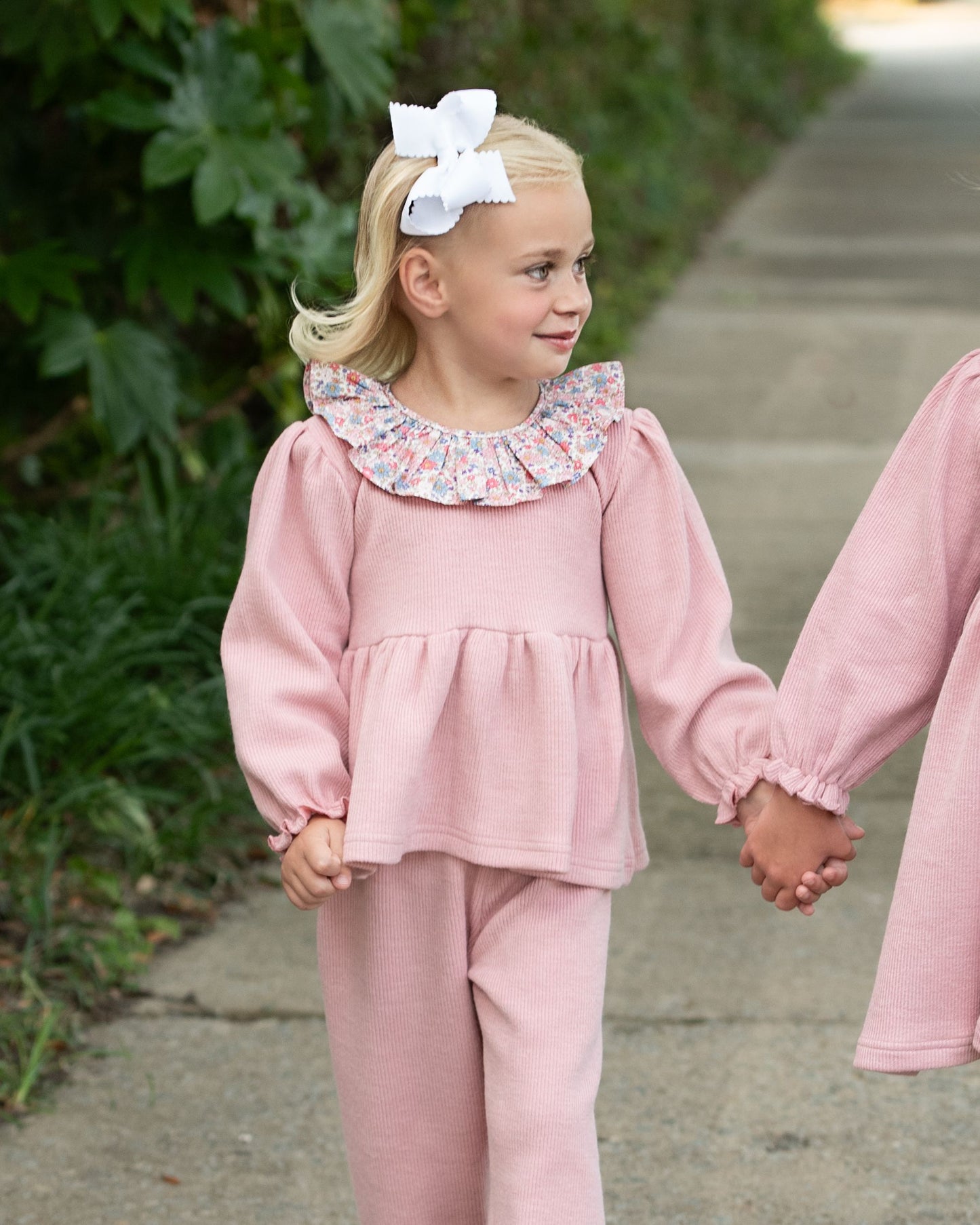 Young girl in a pink outfit with a floral collar holding hands with another person outdoors.