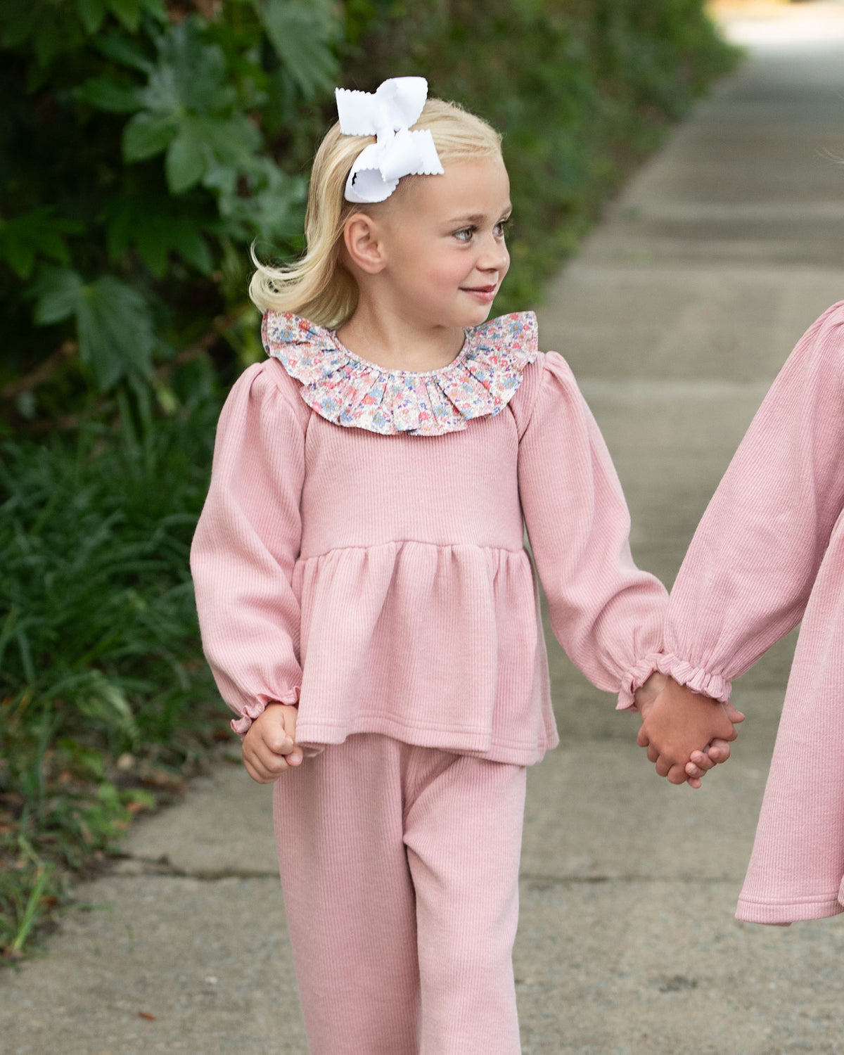 Young_girl_in_a_pink_outfit_with_a_floral_collar_holding_hands_with_another_person_outdoors._1