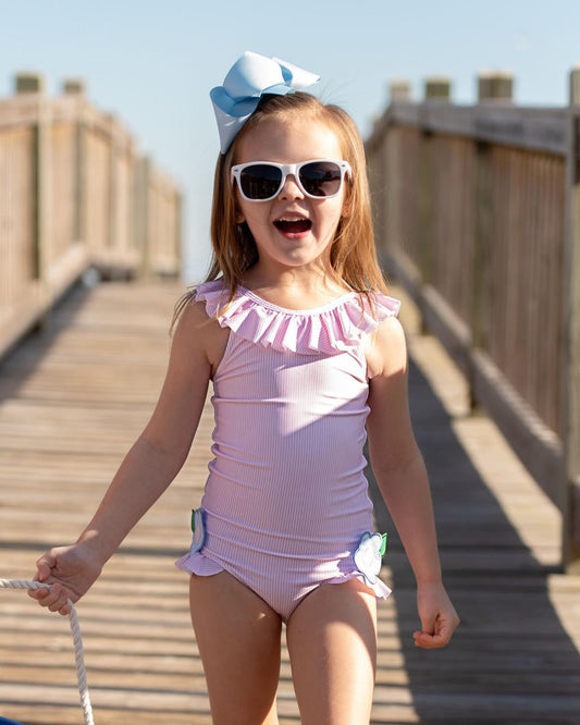 Young girl in a pink swimsuit with sunglasses and a blue bow on a wooden boardwalk.