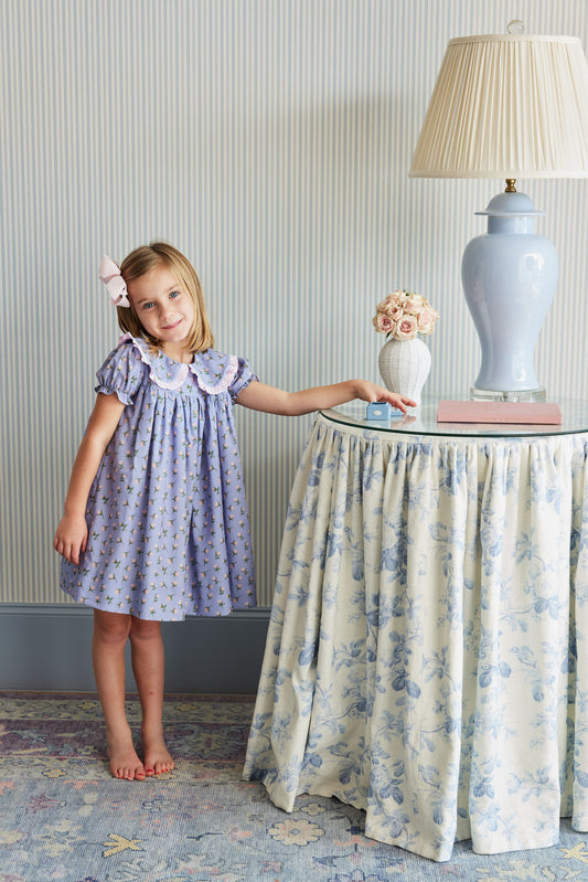 Young girl in a purple dress standing next to a table with decorative items in a room.