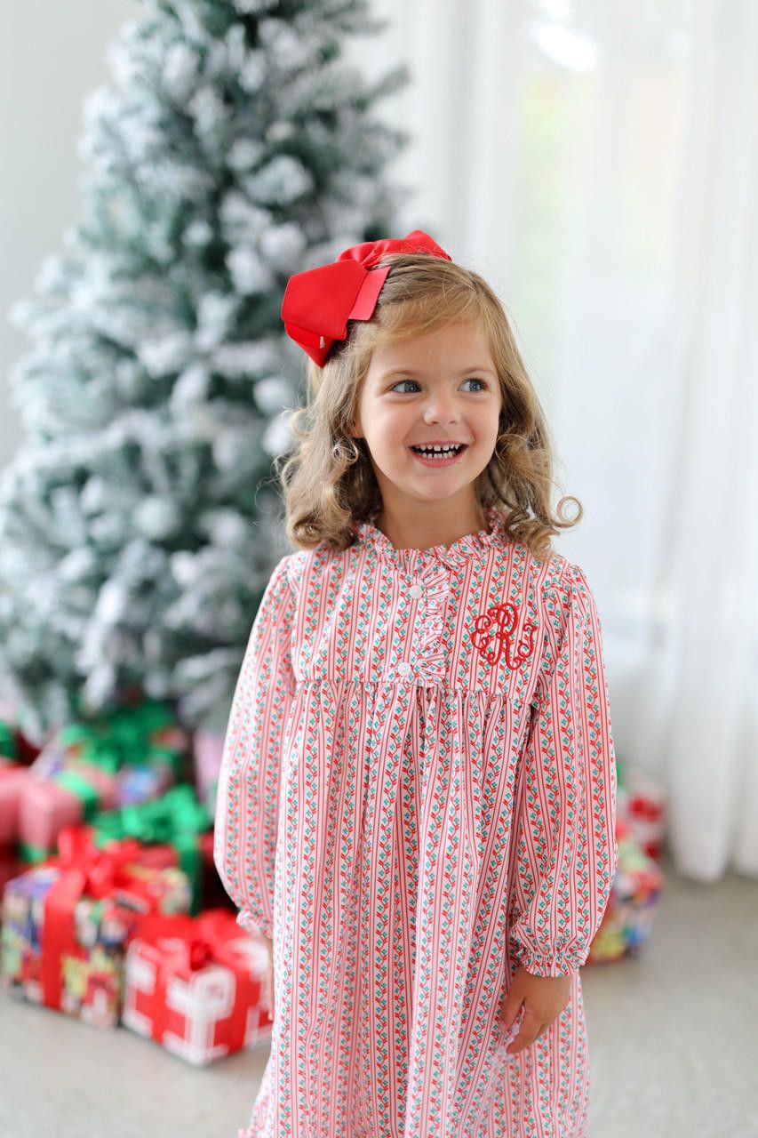 Young girl in a red and white patterned dress with a large red bow standing in front of a Christmas tree and presents.