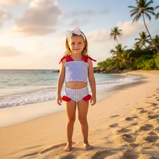 Young girl in a red and white striped swimsuit standing on a sandy beach with palm trees in the background.