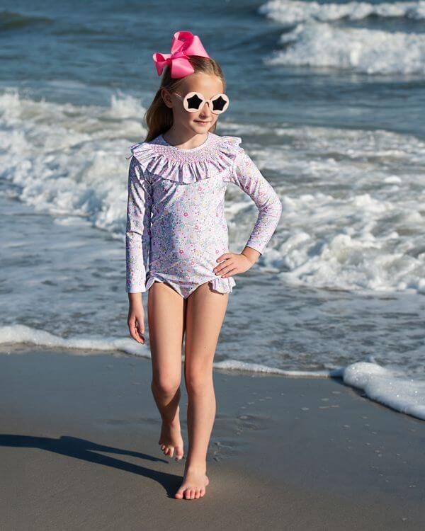 Young girl in a swimsuit and sunglasses standing on a beach with ocean waves in the background