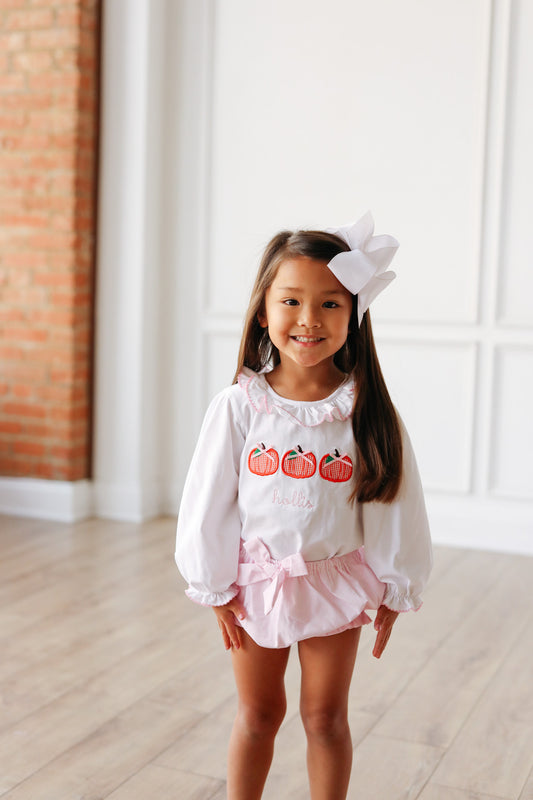 Young girl in a white blouse with pumpkin design and pink shorts standing in a room with brick wall and white floor.