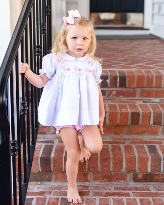 Young girl in a white diaper set with pink trim standing on a set of brick steps.