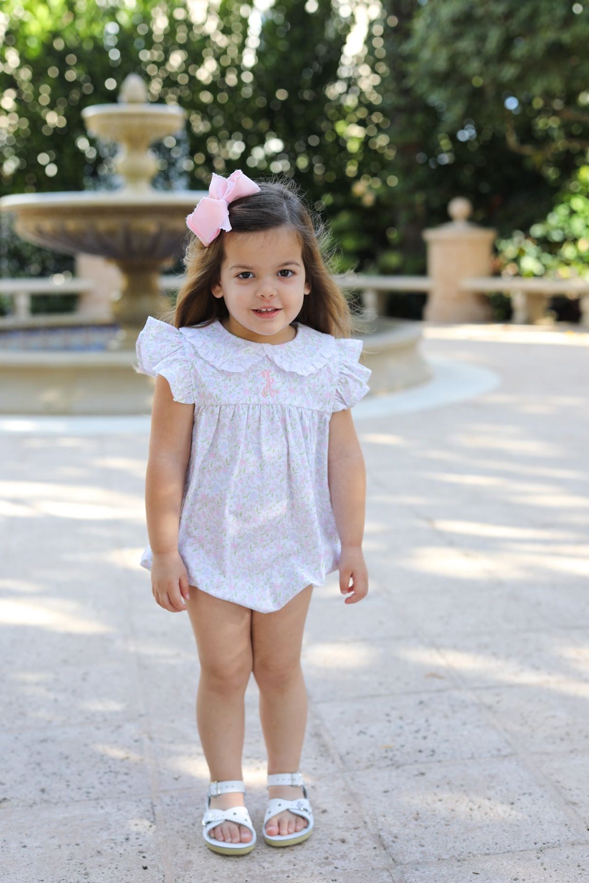 Young girl in a white dress standing in a sunlit outdoor setting with greenery.