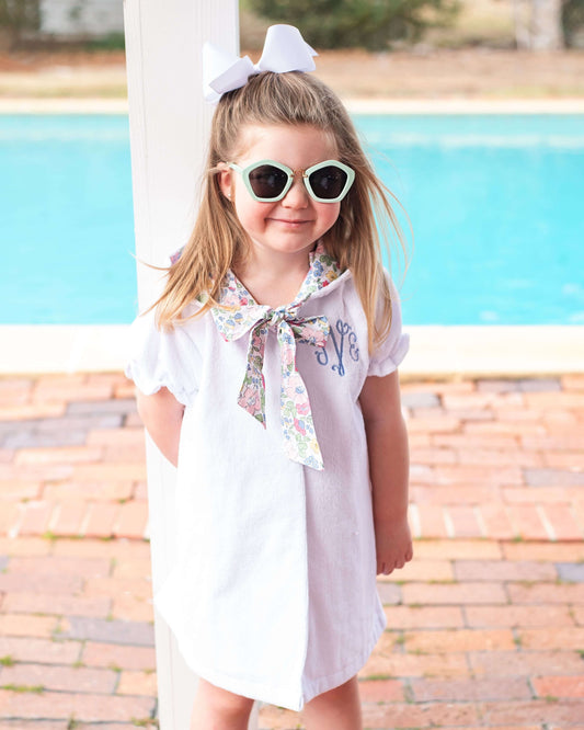 Young girl in a white dress with a floral bow standing by a pool.