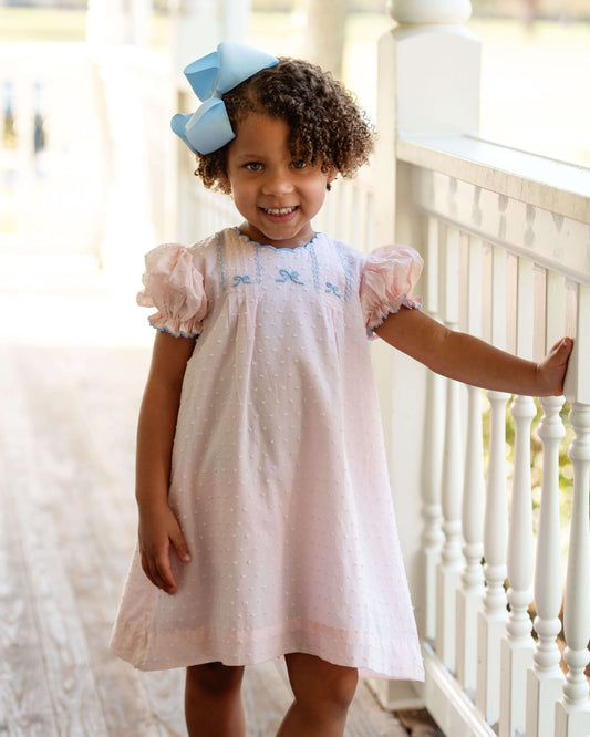 Young girl in a white dress with blue embroidery standing on a porch.