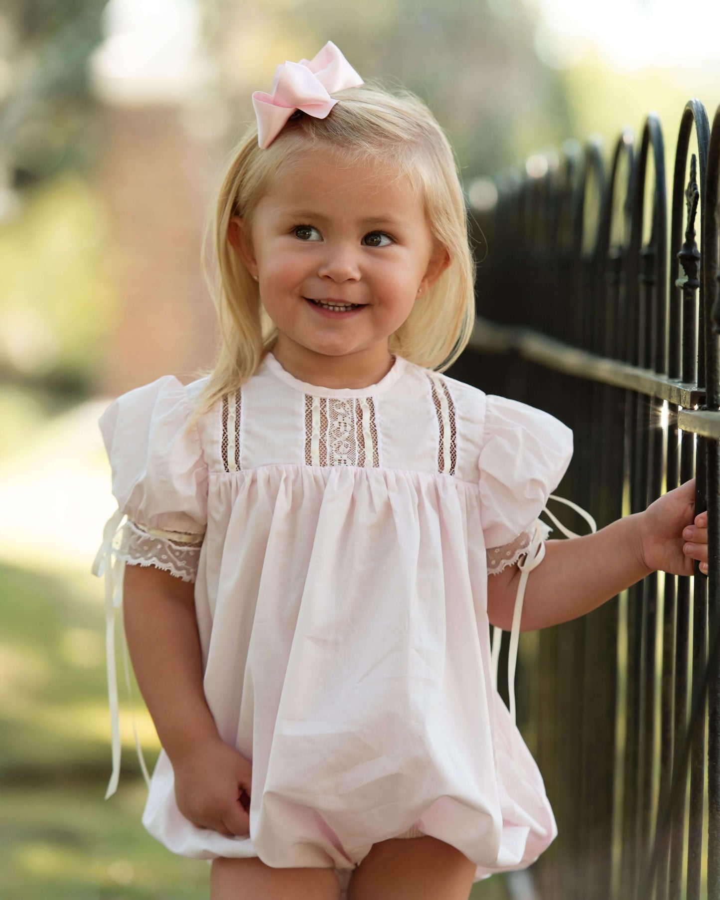 Young girl in a white dress with lace details standing next to a black metal fence.